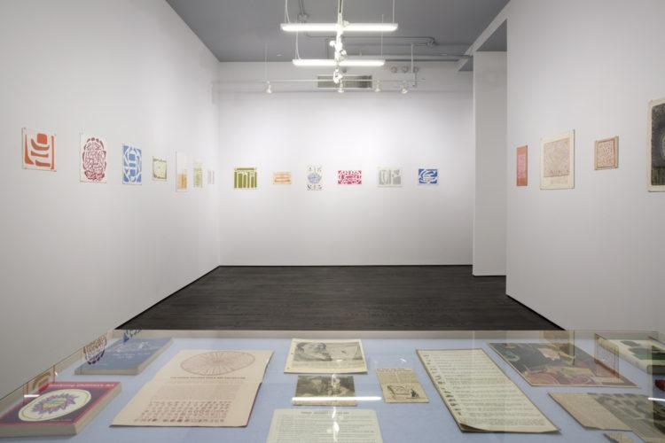Seventeen works installed on white walls in a gallery. All are abstract works on paper. In the foreground, printed ephemera is displayed inside a vitrine table.