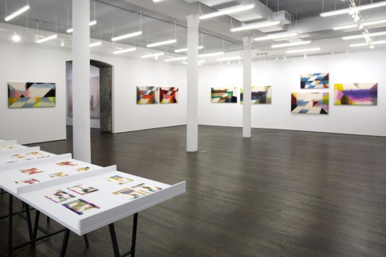 Eight large works installed on walls in a gallery. In the lower left corner, a partial view of a table on which similar, much smaller works are laid out in groups.