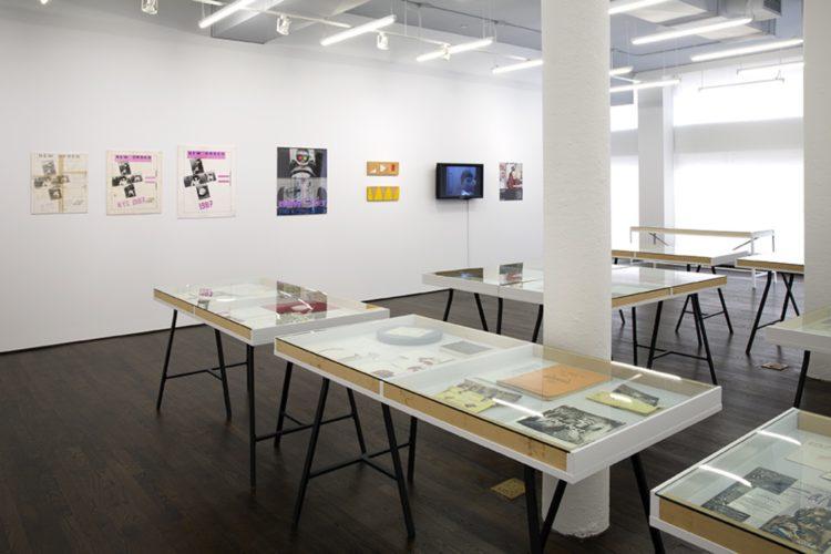 Seven works installed in a row along a wall in the background. In the foreground, several freestanding vitrine display tables are visible.
