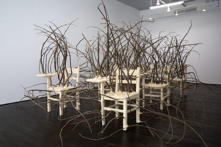 Briar Patch, 2018, a set of six carved wooden school chairs with attached desks. Numerous long, twisting branches extend out of them, nearly engulfing the sculpture.