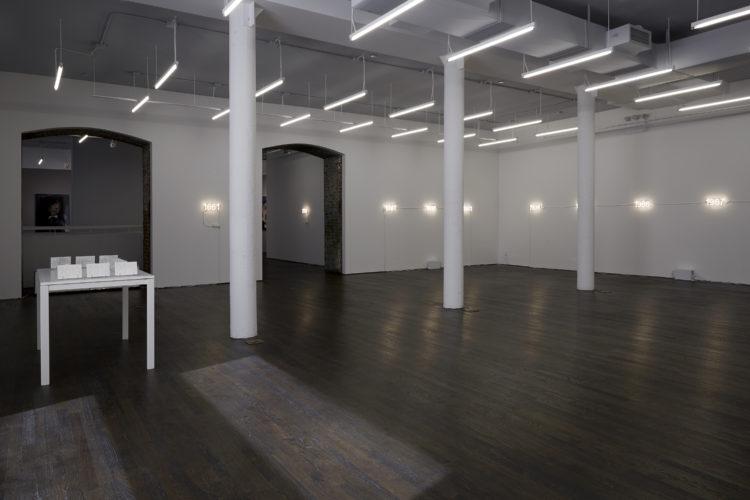 Several illuminated neon signs installed along the walls of a gallery with three columns. The rest of the space is empty except for a table to the left with postcards displayed on stands.